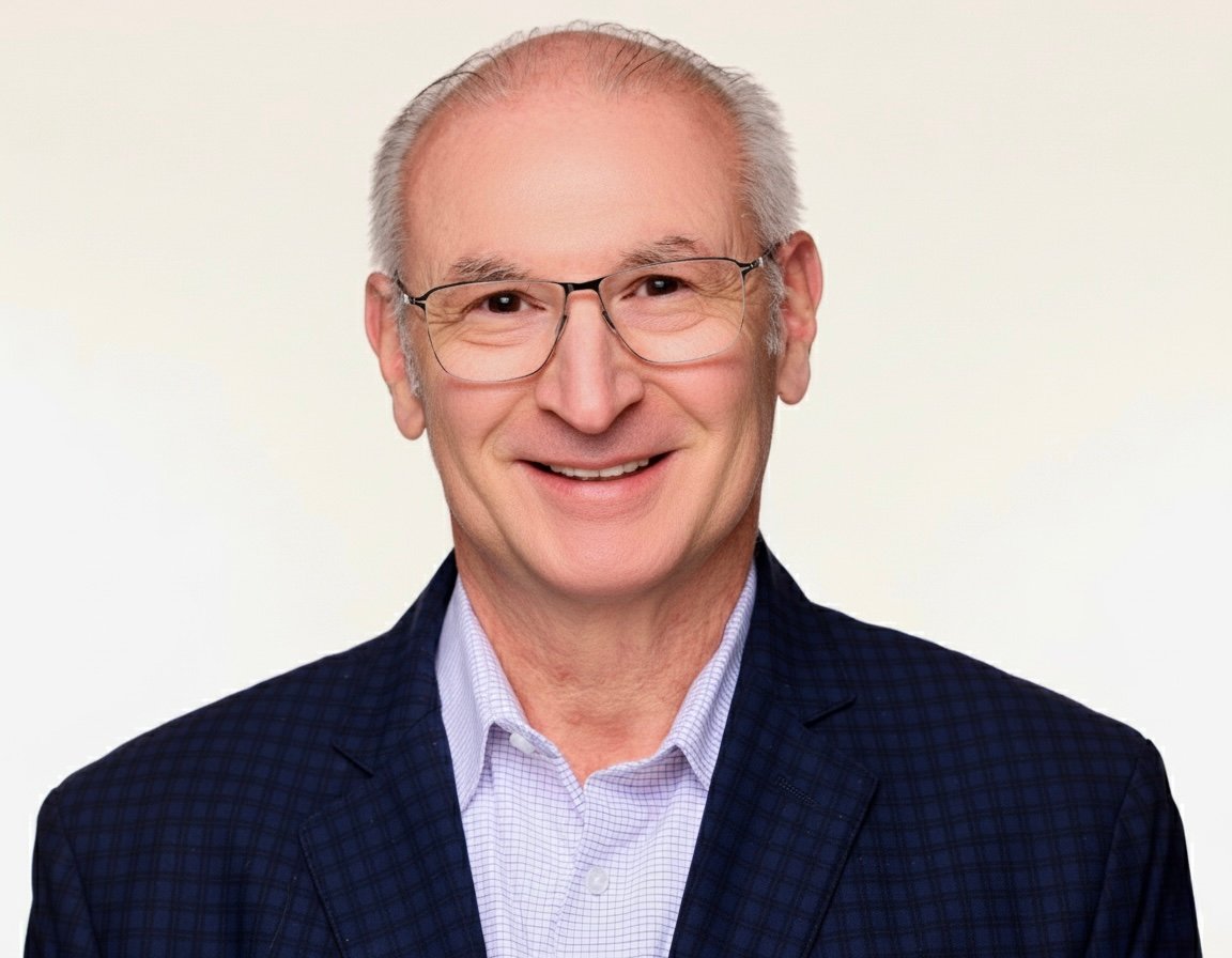 Smiling older man wearing glasses, a navy checked blazer, and a light button-down shirt against an off-white background.