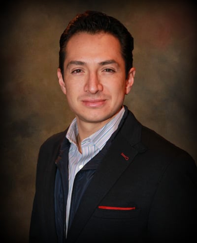 Formal studio portrait of a smiling man in a dark suit with red accents against a mottled brown background.
