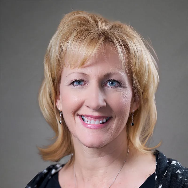 Smiling blonde woman with blue eyes and dangling earrings against a plain gray background.