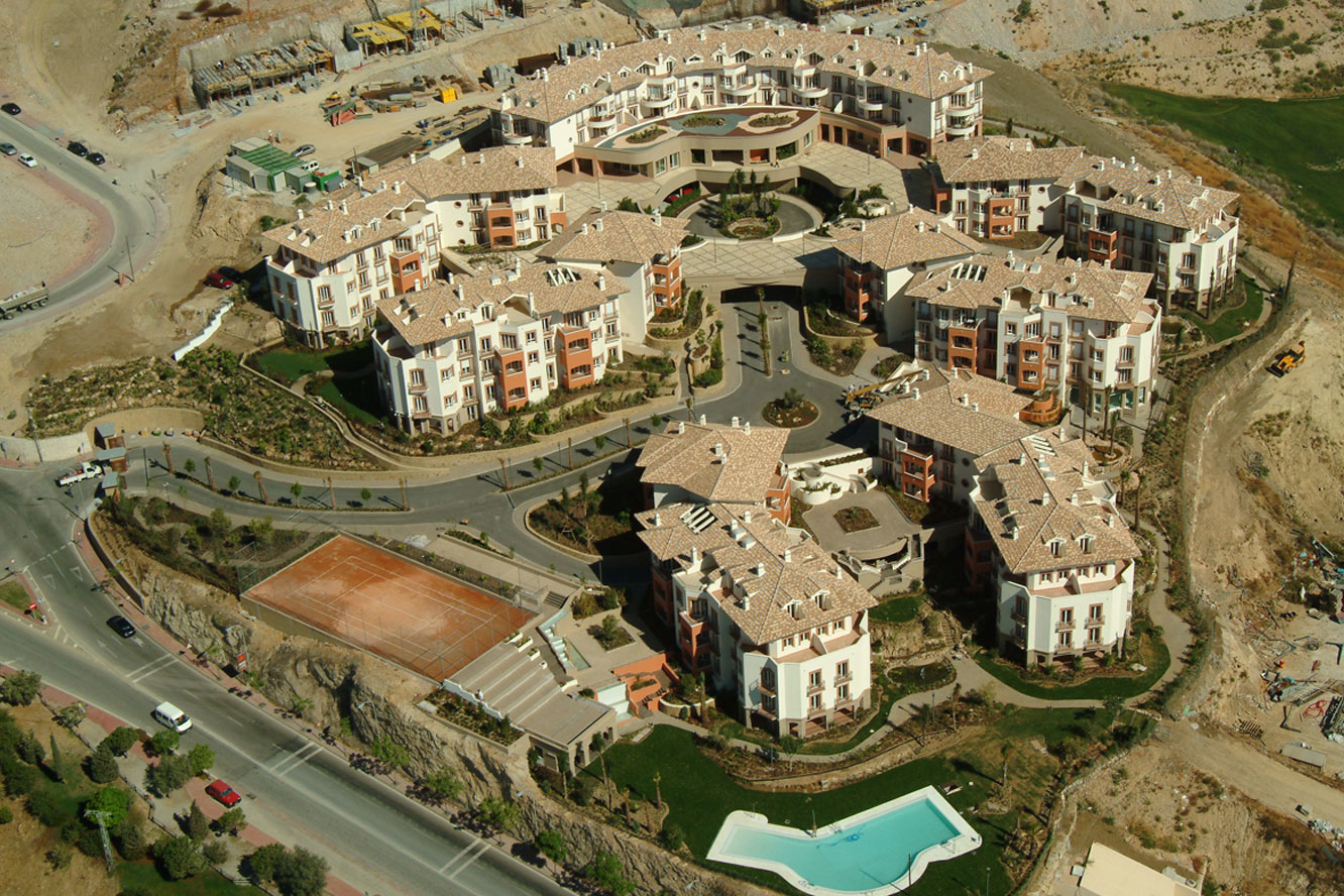 Aerial view of a terracotta-roofed resort complex with courtyards, pools, tennis court, roads, and construction on rocky terrain.